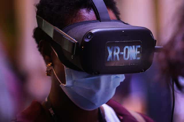 A black woman with short hair wearing virtual reality goggles, a surgical facemask and gold earrings