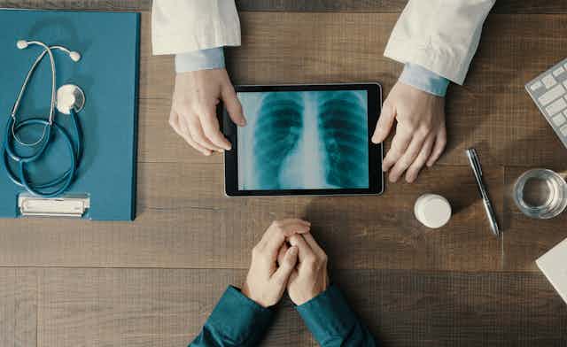 View of a desktop from above. Hands in a white coat on one of the table showing an X-ray, another person's clasped hands are across the desk and a stethoscope lies to the left.