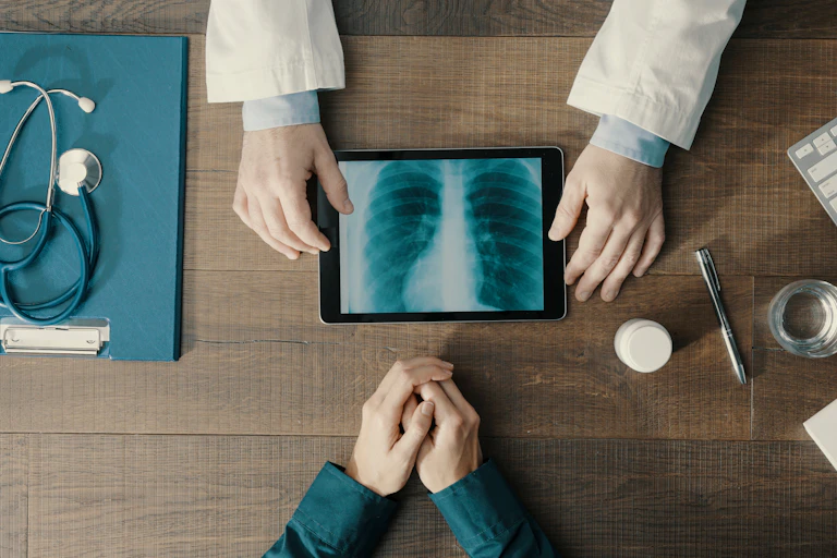 View of a desktop from above. Hands in a white coat on one of the table showing an X-ray, another person's clasped hands are across the desk and a stethoscope lies to the left.