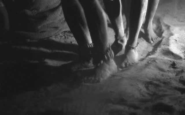 A black and white image of three pairs of feet, dancing in sand, around their ankles are little pods tied to a hide string.