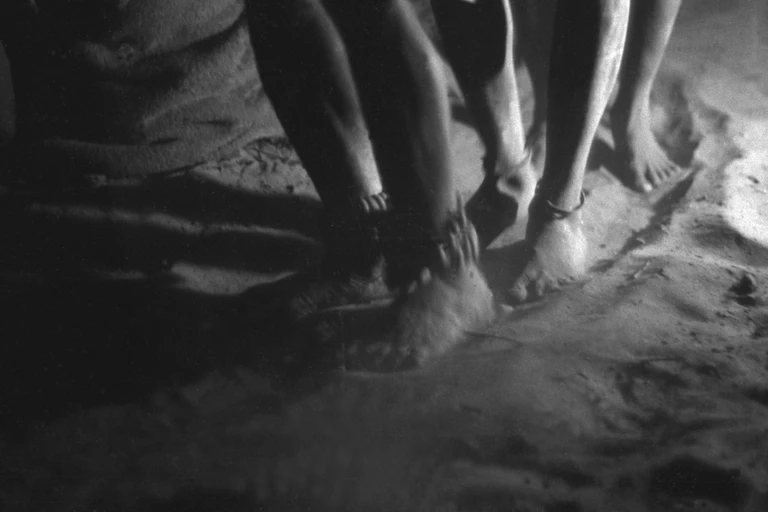 A black and white image of three pairs of feet, dancing in sand, around their ankles are little pods tied to a hide string.