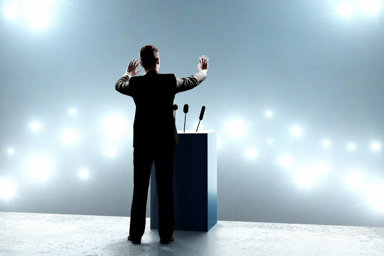 A man in a suit raises his hands at a lecturn on a stage in front of a brightly lit space.