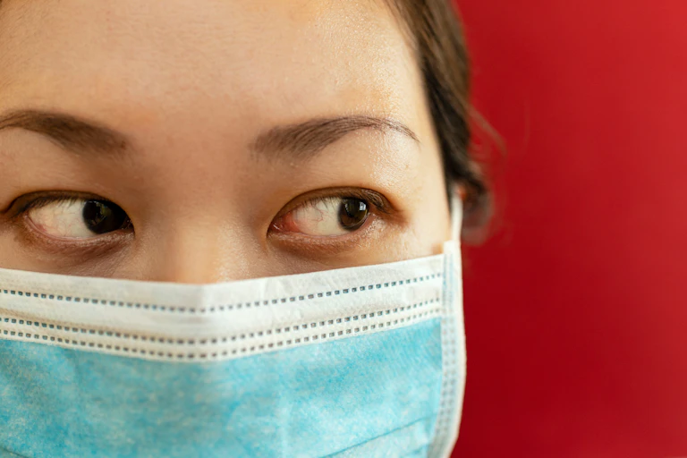 Close up of a woman in a face mask with bloodshot eyes against a red background