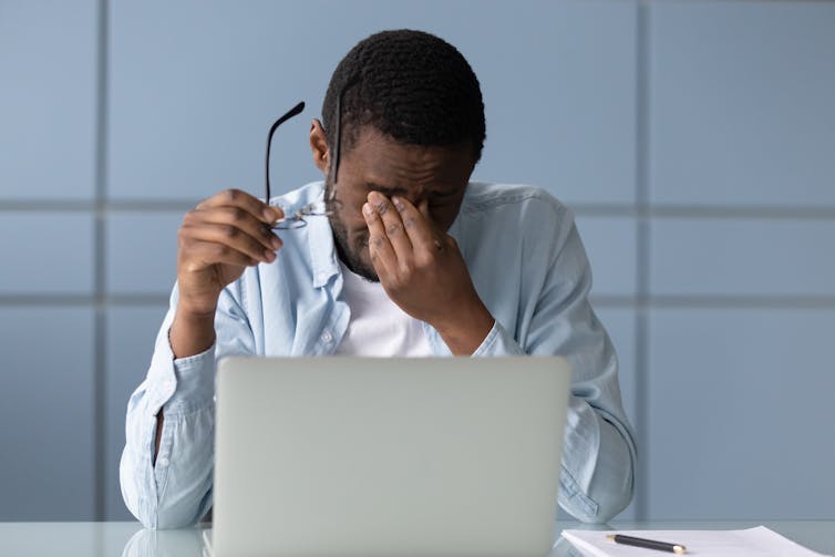 A man sits in front of a laptop, rubbing his eyes with a glass in hand.