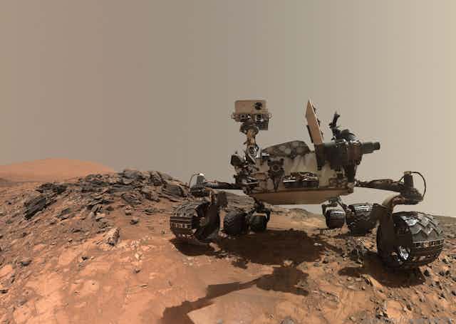 A wheeled robotic rover parked atop reddish brown soil with rocks nearby.