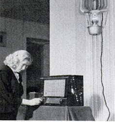 A black and white photo of a woman turning a dial on a large table top radio, with a lantern hanging above it.