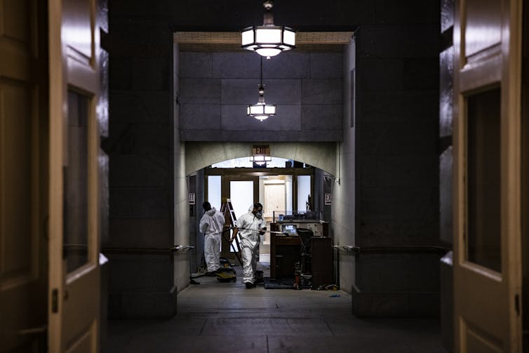 Um corredor escuro no edifício do Capitólio dos Estados Unidos, com trabalhadores em macacões brancos limpando os estragos.