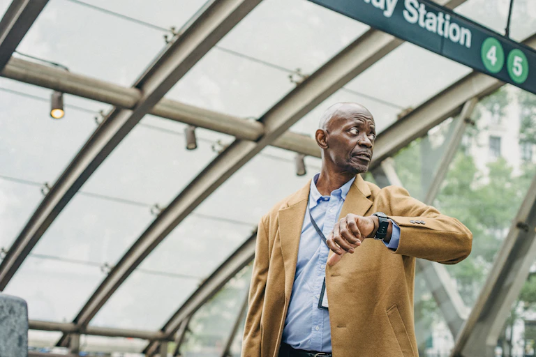 A man in a blue shirt and camel-coloured coat looks at his watch as he leaves a subway station.