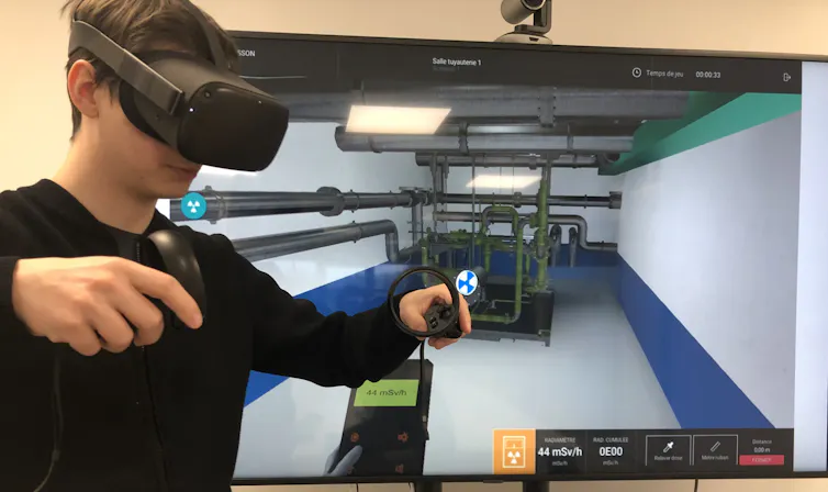 Jeune homme coiffé d’un casque de réalité virtuelles, deux manettes à la main. Derrière lui, un écran montre une installation technique. Un capteur indique la dose de radiation qu’il recevrait en réalité : 44mSv/h