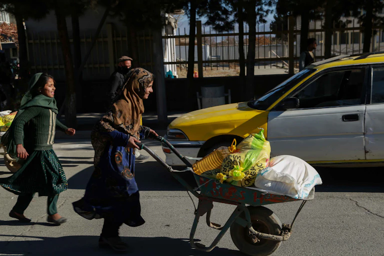 Two Afghan women in traditional dress, one p-ushing a wheelbarrow full of sacks of food supplies.