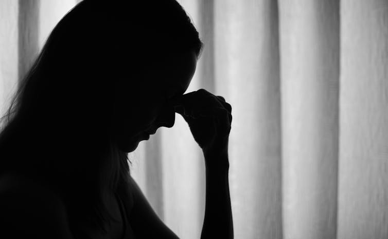 Close up silhouette of a woman looking sad with her hand over her face next to a window