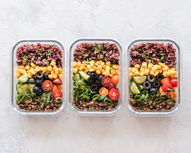 Plant based meal prep, three tupperware containers are shown with rice, corn, avocado, tomato, beans and olives