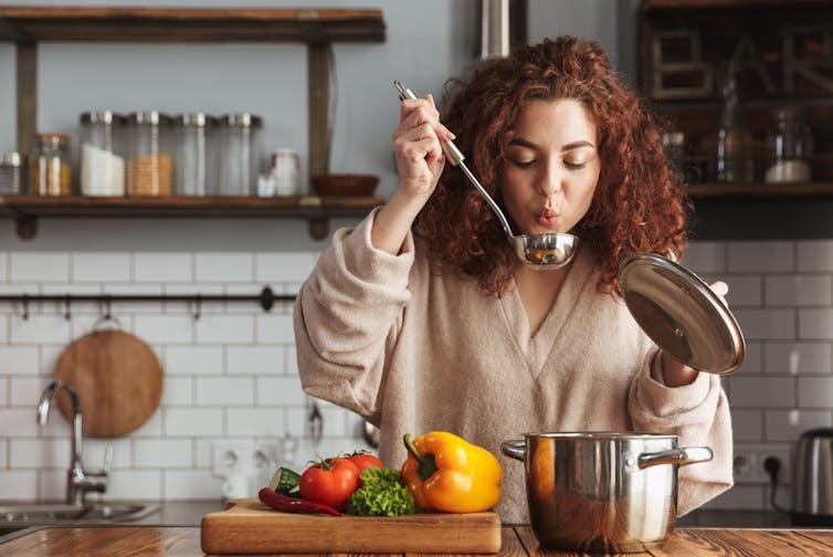 A woman blows into a ladle as she lifts it out of a pot of soup, vegetables are on a cutting board in front of her
