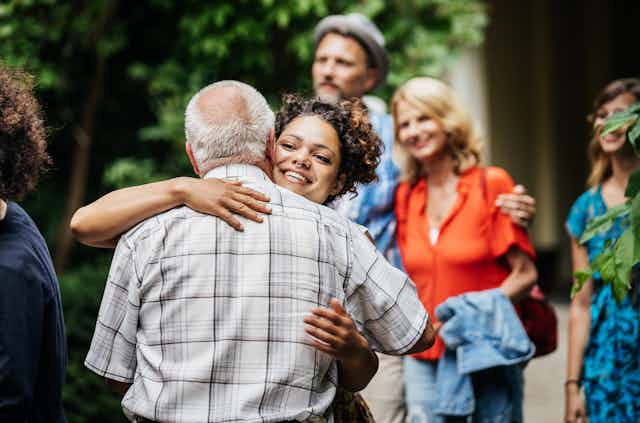 Elderly man hugging young woman outdoors, with middle-aged family members in background.