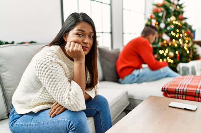 A young couple with glum expressions, sitting apart on a sofa, with a Christmas tree in the background.
