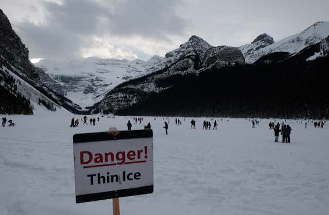 A 'Danger! Thin Ice' sign is placed in Alberta's Lake Louise, with people skating in the background.