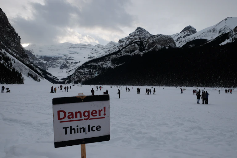 A 'Danger! Thin Ice' sign is placed in Alberta's Lake Louise, with people skating in the background.