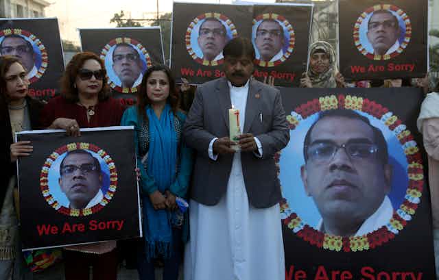 Un hombre vestido con una chaqueta y una camisa larga blanca sostiene una vela, mientras dos mujeres de pie junto a su casa sostienen la fotografía de un hombre que fue linchado en Pakistán.