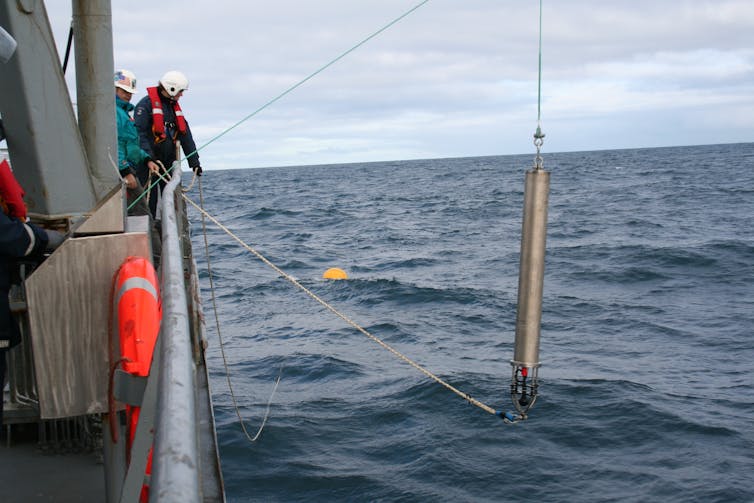 Two people wearing life jackets on a boat lower a long, metal cylinder into the sea.