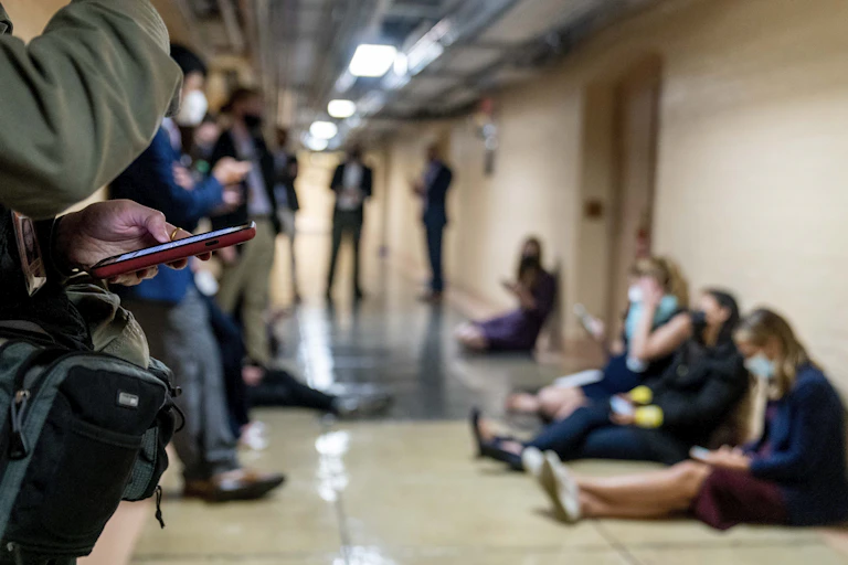 Reporters in a hallway, sitting on the floor and standing outside a meeting room.