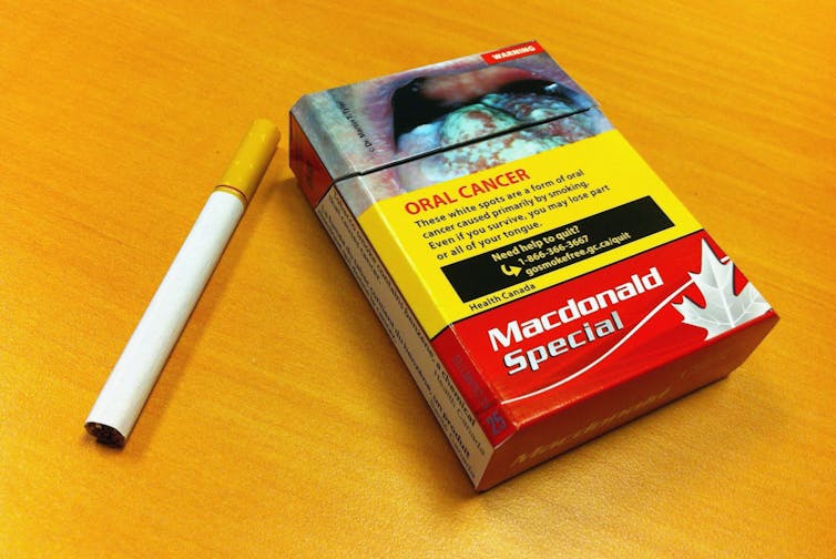A package of cigarettes on a table with one cigarette beside it. The box shows a photograph of a tongue covered in white spots, a form of oral cancer caused primarily by smoking.