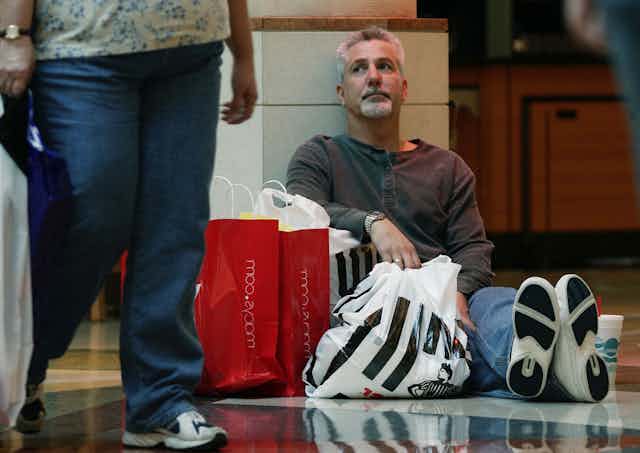 Man sits on mall floor surrounded by shopping bags.