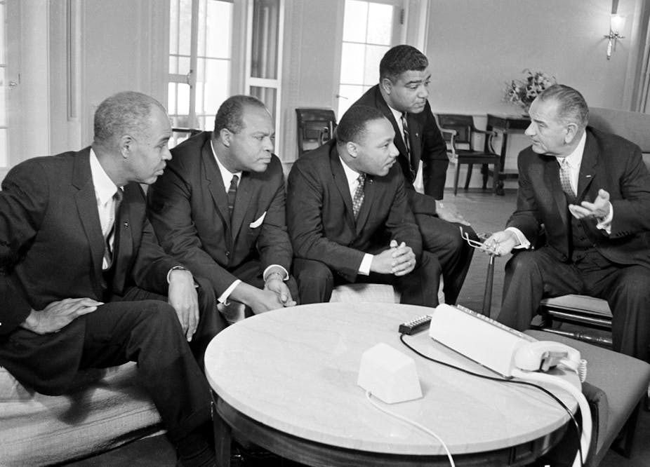 Five men dressed in black suits, including President Lyndon B. Johnson and Martin Luther King Jr., holding a discussion.