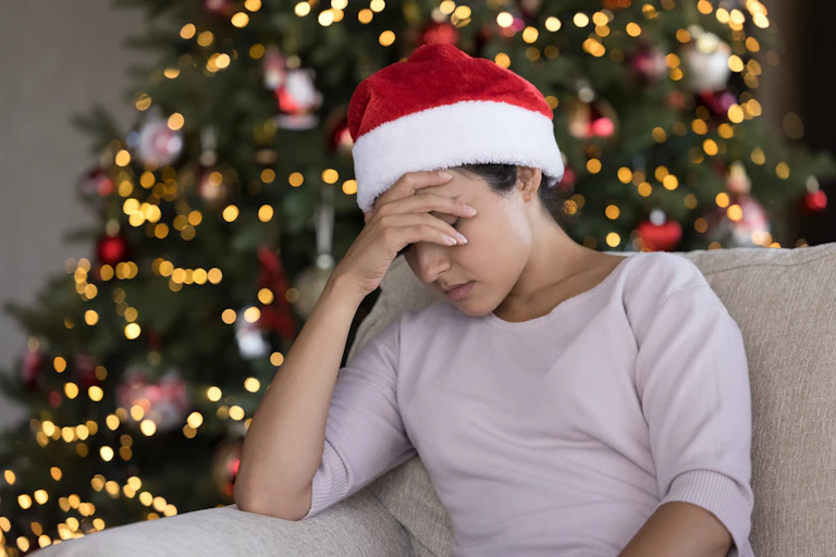 A young woman wearing a santa hat with her head in her hand in front of a Christmas tree