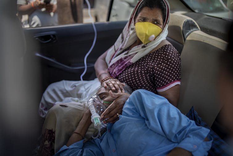 A man wearing an oxygen mask rests his head on a woman's lap. Both are in the backseat of a car.