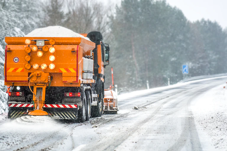 A deicer clears ice from a road lined by trees and showers road salt to melt the ice.