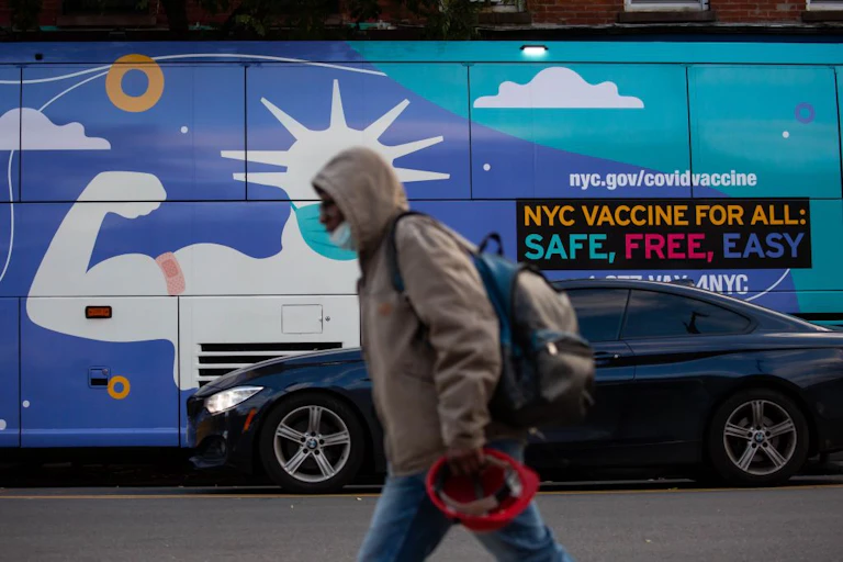 A pedestrian walks in front of a van with an ad that says "NYC VACCINE FOR ALL: SAFE, FREE, EASY"