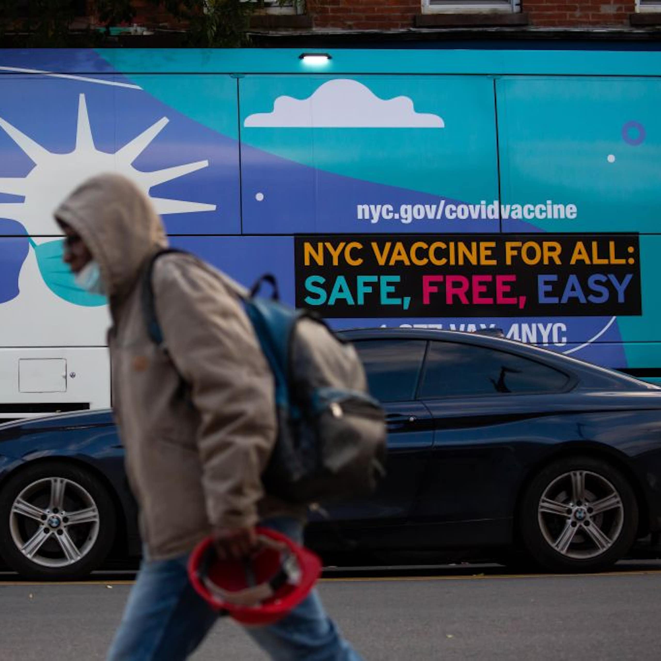 A pedestrian walks in front of a van with an ad that says "NYC VACCINE FOR ALL: SAFE, FREE, EASY"  