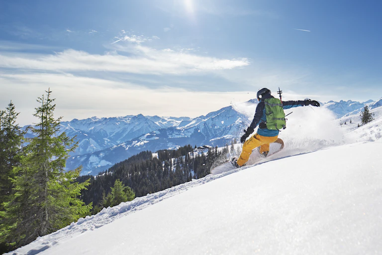Snowboarder carving a line down a mountain in fresh powder with trees, mountains and sky in the background.