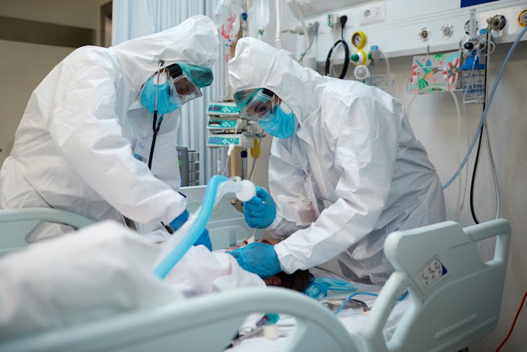 Two doctors leaning over a patient while intubating, or placing the patient on a ventilator.