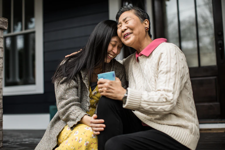 A younger woman and older woman sit together gently laughing on porch steps.