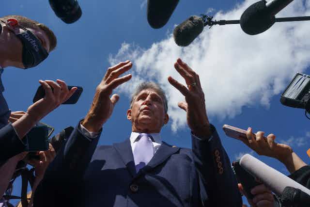 Senator Manchin gesticulates with both hands as a swarm of boom mics surround him during an interview, with the sky in the background.