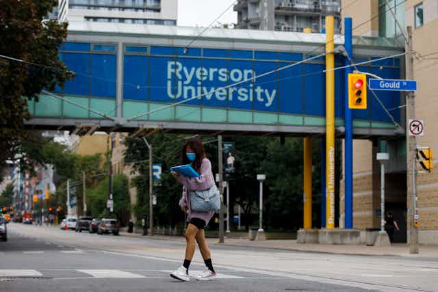 A girl wearing a mask crosses Gould St. in Toronto. Ryerson University is displayed on a walkway behind her.
