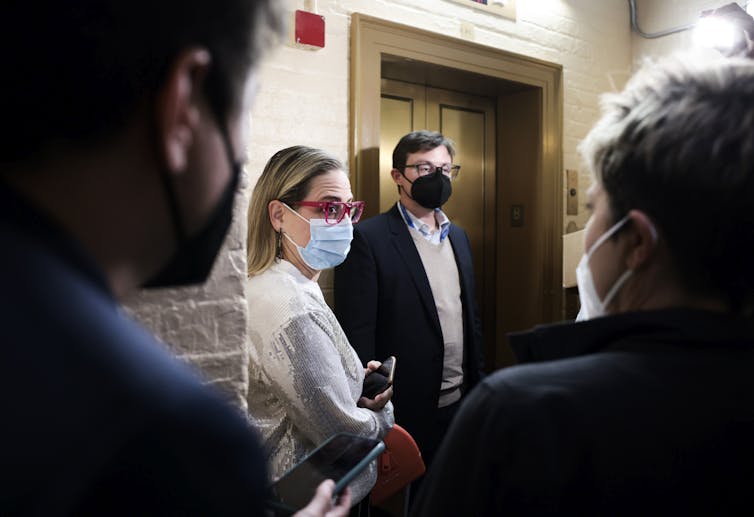 Sen. Kyrsten Sinema, masked, leaving an office in the Senate, surrounded by people.