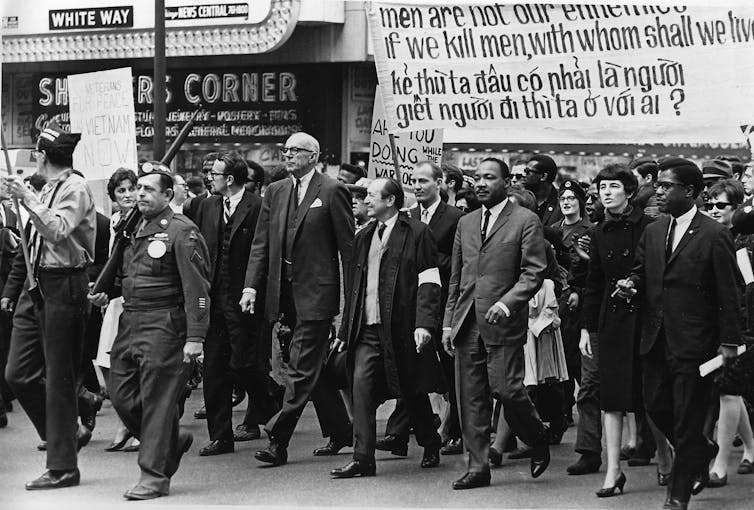 Led by Martin Luther King Jr., several men dressed in black suits march in a rally. Ahead of them are police officers holding rifles.