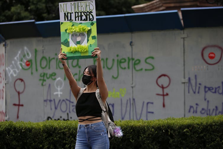 A woman holds a placard that reads in Spanish 'Neither Dead Nor or in Prison,' with graffiti behind her