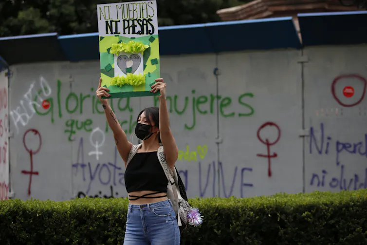 A woman holds a placard that reads in Spanish 'Neither Dead Nor or in Prison,' with grafitti behind her