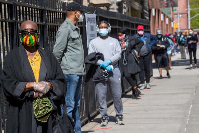 A line of people, all wearing masks, wait in line on a sidewalk