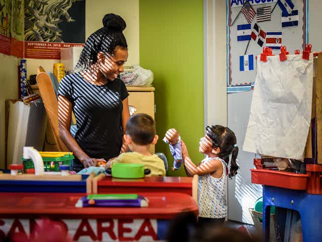 A Black female child care worker plays with two small children in a classroom.