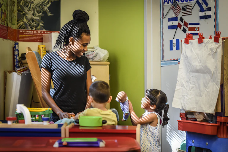 A Black female child care worker plays with two small children in a classroom.