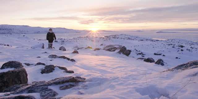 A woman is seen in a parka against an arctic snowy landscape and the sun at the horizon.