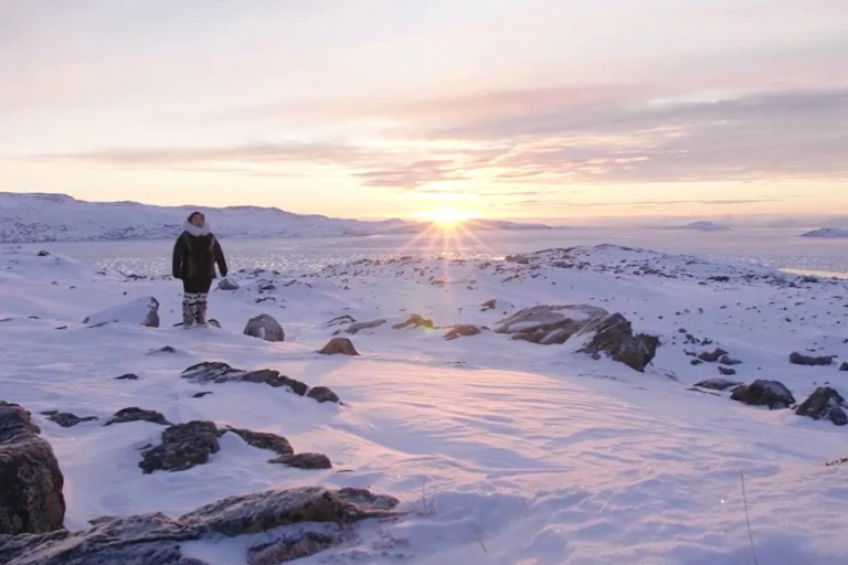 A woman is seen in a parka against an arctic snowy landscape and the sun at the horizon.