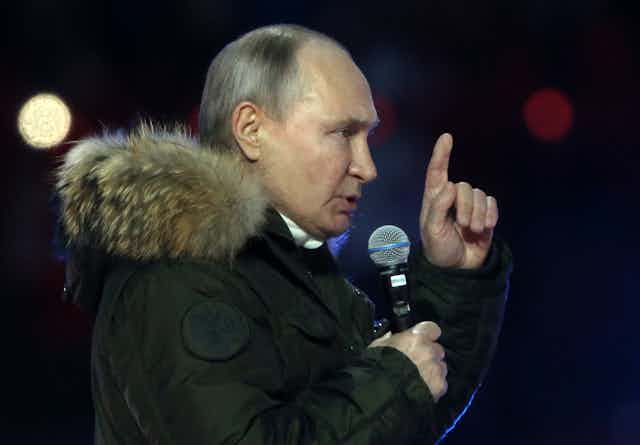 Russian President Vladimir Putin gestures with his hand as he talks into a microphone while wearing a thick winter coat.