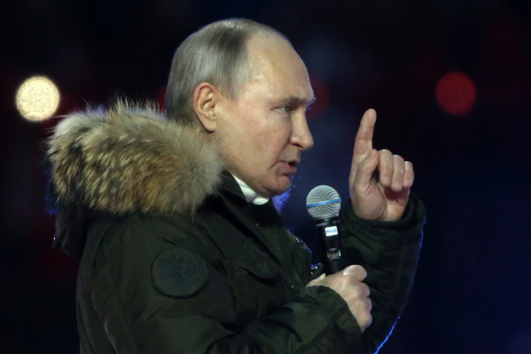 Russian President Vladimir Putin gestures with his hand as he talks into a microphone while wearing a thick winter coat.