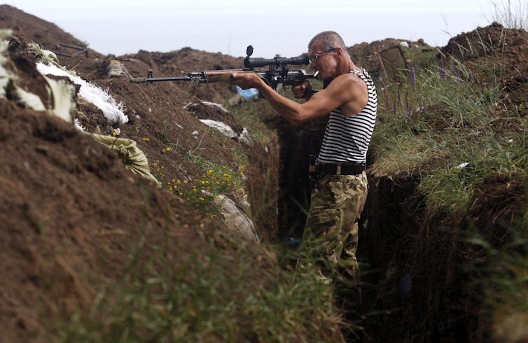 A soldier stands in a trench while looking down the scope of a rifle.