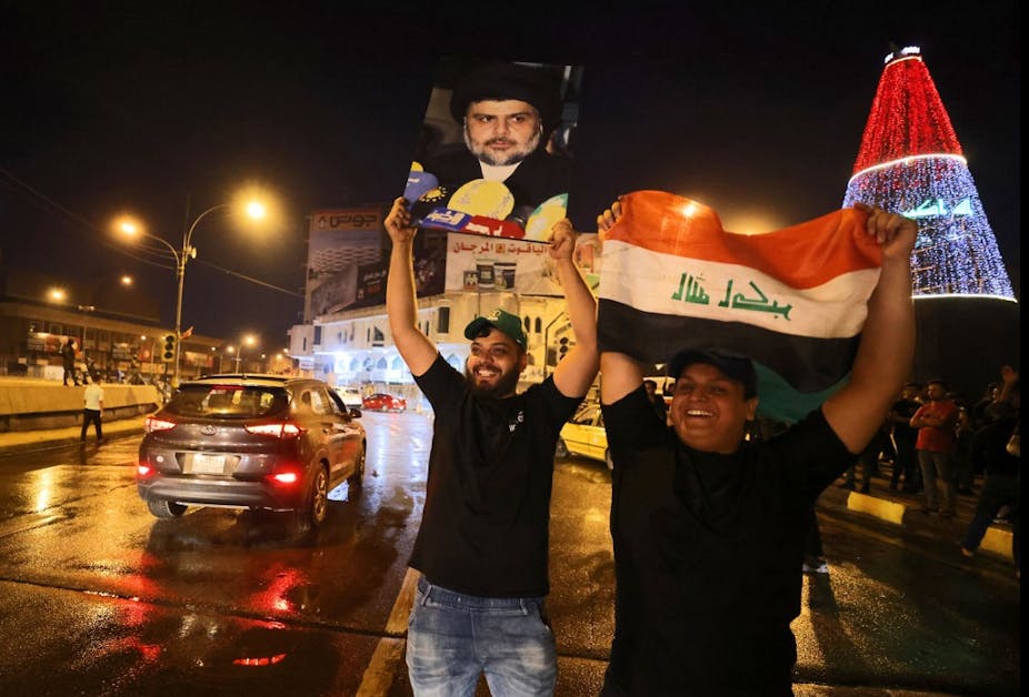Deux hommes brandissent un drapeau irakien et un portrait de leader politique au bord d'une route en souriant.
